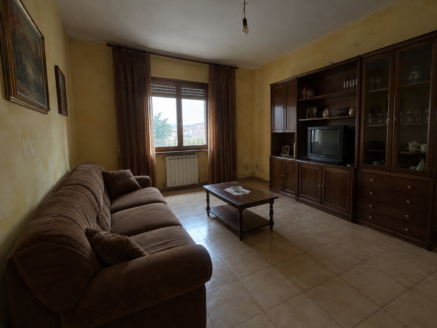 Esempio inventato: soggiorno datato con pareti gialle macchiate, piastrelle beige, divano marrone, parete attrezzata scura e televisore a tubo